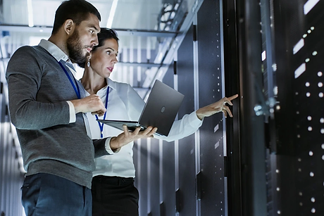 Two IT professionals stand in a server room. One holds a laptop while the other points at a server rack, both focused on the equipment. The background shows rows of servers and blue lights.