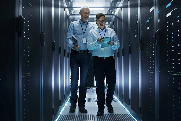 Two men wearing lanyards walk through a server room lined with computer racks. One holds a tablet, and the other holds papers. The environment is modern and has blue-toned lighting.