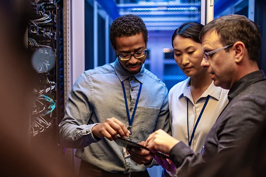 Three IT professionals stand in a server room, examining a digital tablet together. They appear focused and are surrounded by server racks and blue lighting.