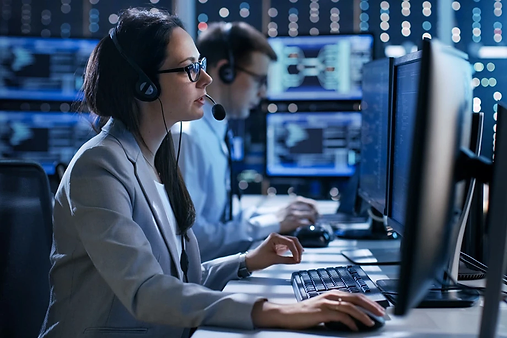 A woman wearing a headset works at a computer in an office with multiple monitors. Another person is seated and working in the background. The setting appears to be a control room or call center with dim lighting.