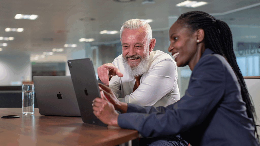 A pair of business people engaged in discussion at a table with a Mac and Ipad open between them.
