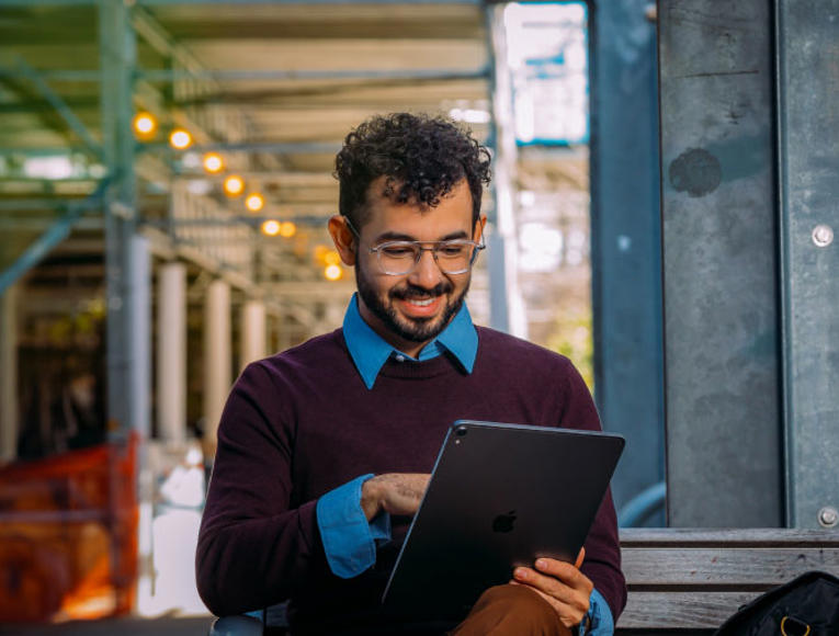 A man wearing glasses and a sweater sits on a bench, focused on using an iPad.