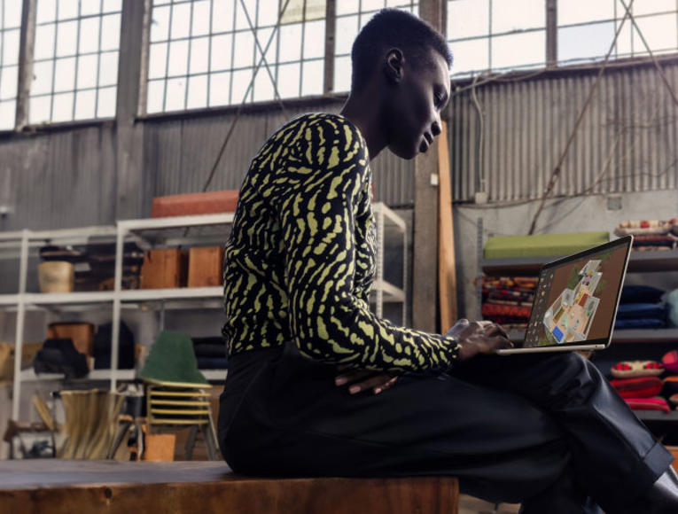 A man sitting on a bench, focused on his laptop, in a indoor office in the background.